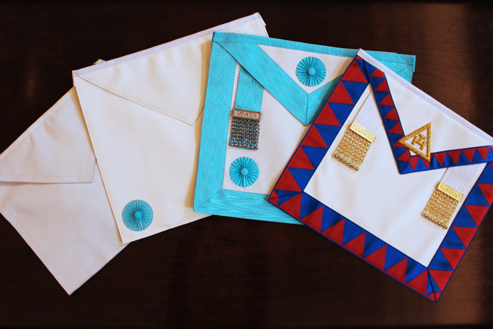 Four Masonic aprons displayed in a fan arrangement, showing the progression from plain white through to the decorated Royal Arch apron